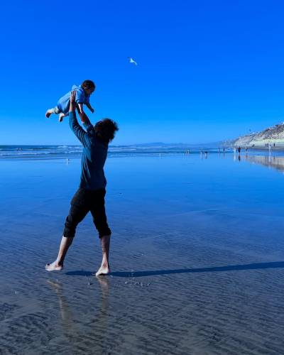 Photo of Samba seen from the side as he lifts his kid up above his head. He is looking up at them, but his face is hidden behind his hair. The photo is shot in the same beach setting as the previous picture.