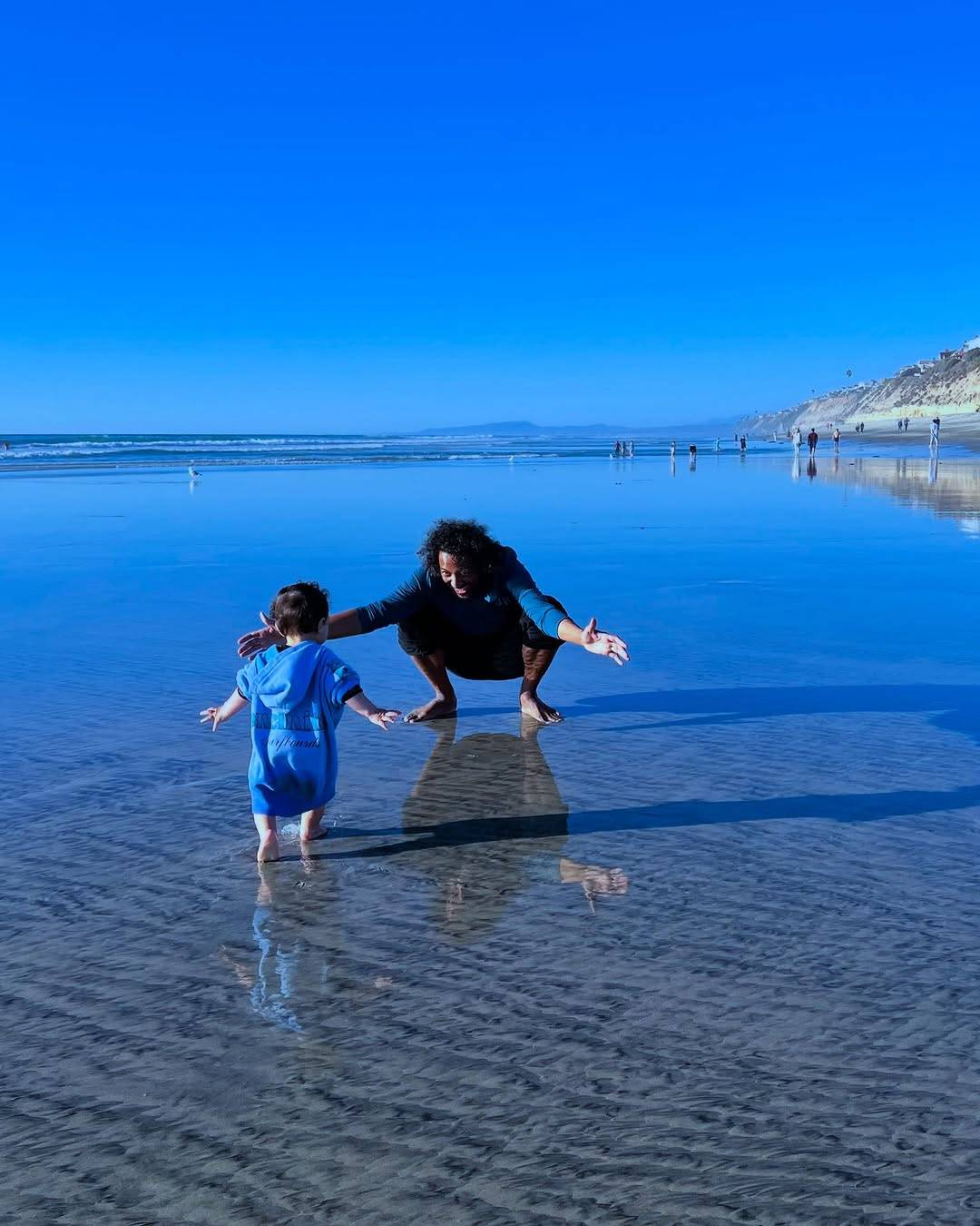 Another photo from the same set. Samba squats down in the wet sand with his arms still outstretched and a big smile on his face as his kid runs towards him.