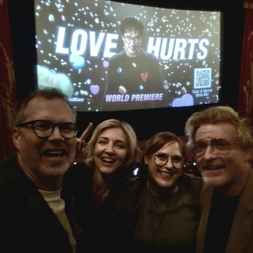 Photo from the Love Hurts premiere: Rhys and his wife Rosie, another woman and his friend Buttons in front of a large screen, inside a dimly lit movie theatre. They are standing close together and smiling at the camera, their faces looking a bit blurry because of the dim light. The screen behind them displays the movie's title and an image of its lead Ke Huy Quan striding towards the camera with many candy hearts falling down on him, and the words 'world premiere' written at the bottom.