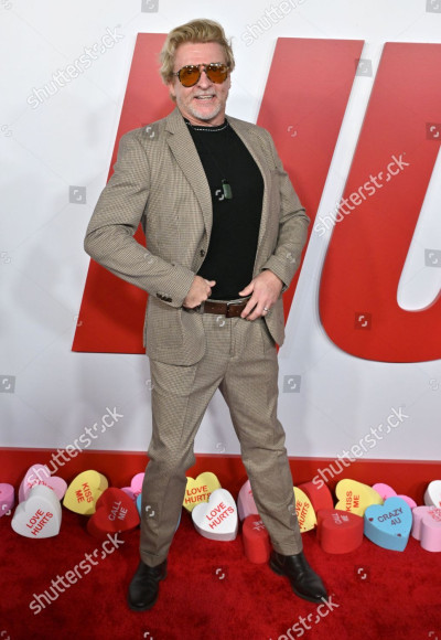 Rhys Darby poses with his hands on his belt and his chest puffed out slightly on the red carpet at the Love Hurts premiere. He is wearing a tan suit with a fine houndstooth pattern, black shoes and a black t-shirt, paired with a pounamu stone necklace and aviator sunglasses.