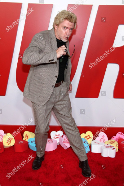 Another photo of Rhys posing on the red carpet, with his upper body and face slightly turned to the side. He has taken off his sunglasses and is looking sideways at the camera, bringing the end of one of his glasses' arms up between his pouted lips.