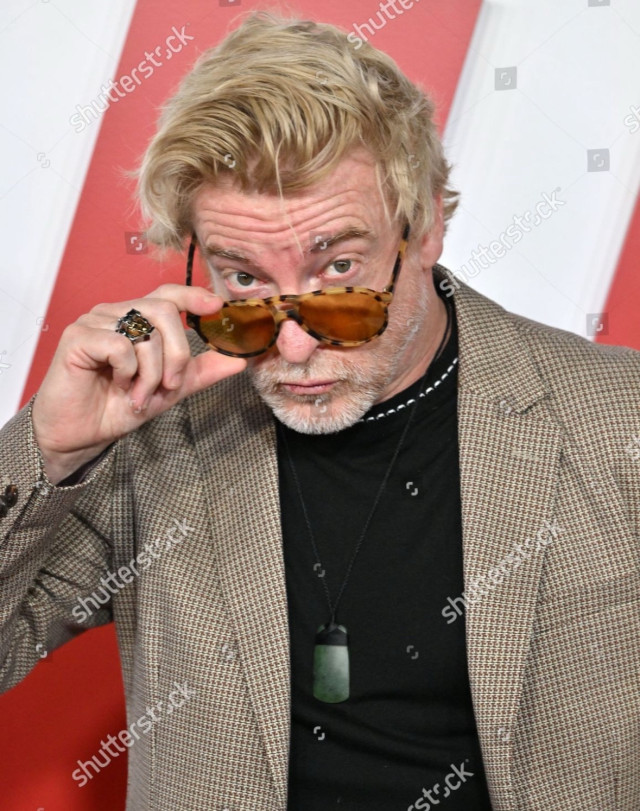 A close-up of Rhys, seen from about the waist up, on the red carpet. With a slight pout and creased brow, he looks at the camera over the rim of his sunglasses, which he has pulled down his nose a little with the thumb and forefinger of his right hand. He is wearing chunky ring with a large stone on his ring finger.