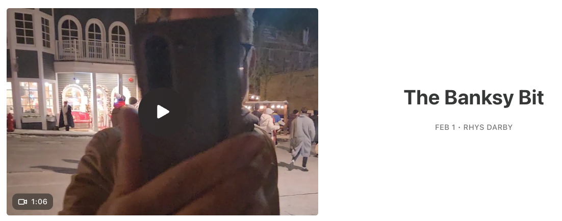 Screenshot of a video posted on Rhys Darby's Substack. The thumbnail shows a phone held right up to the camera by his friend Buttons, with lit-up shop windows and passers-by seen in the background. Next to the video, 'The Banksy Bit - Feb 1, Rhys Darby' is written in black on a white background.