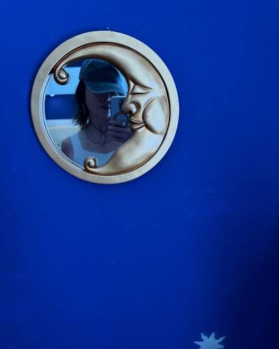 A round mirror with the shape of a crescent moon nestled into its golden frame, hung on a midnight blue wall. The moon has closed eyes, a nose and a smiling mouth. Reflected in the mirror, we see part of Ruibo's face and one of her shoulders. The phone she is taking this photo with obscures one half of her face, and a baseball cap's visor covers her eyes. At the bottom edge of the photo, we can see the top half of a silver star spray-painted onto the wall.