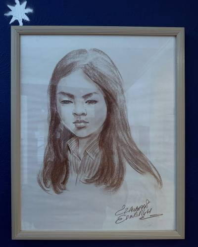 Photo of a framed charcoal drawing of a young girl, possibly Ruibo as a kid, hung on a midnight-blue wall. Her head is turned just slightly to the side and her expression is neutral, her features relaxed and her straight dark hair falling to just below her shoulders. A silver eight-pronged star has been spray-painted onto the wall just above the top left corner of the picture frame.