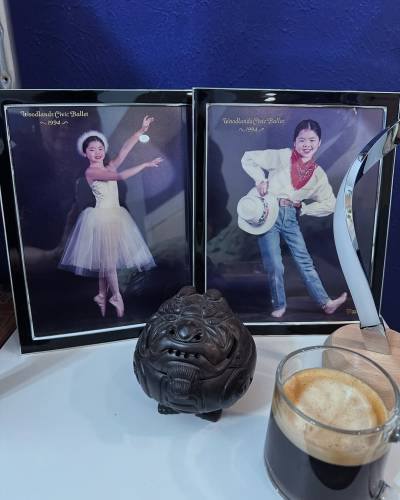 Two framed photos stand side by side on a side table. Both of them show a young girl and have 'Woodlands Civic Ballet - 1994' written in curly gold letters at the top. The photo on the left features a girl in a white tutu smiling at the camera as she stands en pointe with both arms extended to one side, her left arm raised slightly higher than the right. Her black hair is tied back and a fluffy white headpiece circles the back of her head. The photo on the right shows a girl in a cowboy costume, smiling as she stands with her upper body titled to the left and both hands on her hips, one hand holding a white cowboy hat. She is wearing a red neckerchief, a white blouse and blue jeans.