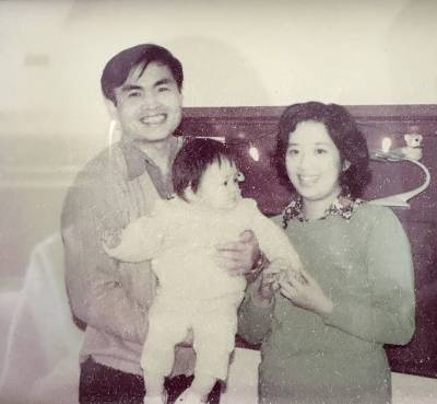 A black & white photo showing a smiling man and woman, the man holding a baby who is looking at something off to the side. The woman is holding one of he baby's hands in both of her own.