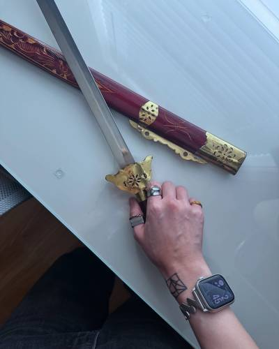 Photo showing part of a narrow sword or long knife and a wooden sheath lying on a shiny white surface, with Ruibo's hand around the handle. The crossguard is a kind of scalloped trapezoid shape, made from what looks like thin, golden metal with a cut-out flower shape at the center. The sheath is made from lacquered red wood with ornamental details carved into it. Thin golden sheets of metal with ornamental cut-outs similar to those on the crossguard are wrapped around the bottom part of the sheath near its openig.