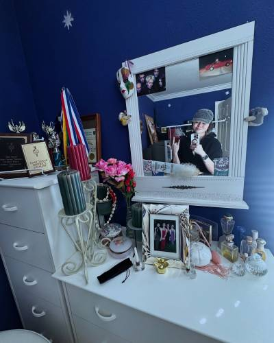 Selfie of Ruibo, seen from the waist up in a wall mirror hanging on a midnight blue wall. She is making devil horns with one hand and holding up her phone with the other, looking down at the phone screen with a smile. The mirror has a thick, white, kind of neo-classical frame, and it hangs above a white dresser, on top of which there are various knick-knacks, perfume bottles, candles and a framed photo of 4 girls in prom dresses. Several medals, trophy statues and framed certificates are displayed on a taller white dresser next to it. The mirror itself is also decorated, with 2 little koalas (?) clipped to the outer edge of the frame on either side, a mid-90s photo of Radiohead along with other pictures tucked into the corners of the mirror, and a masquerade mask hanging from one corner.