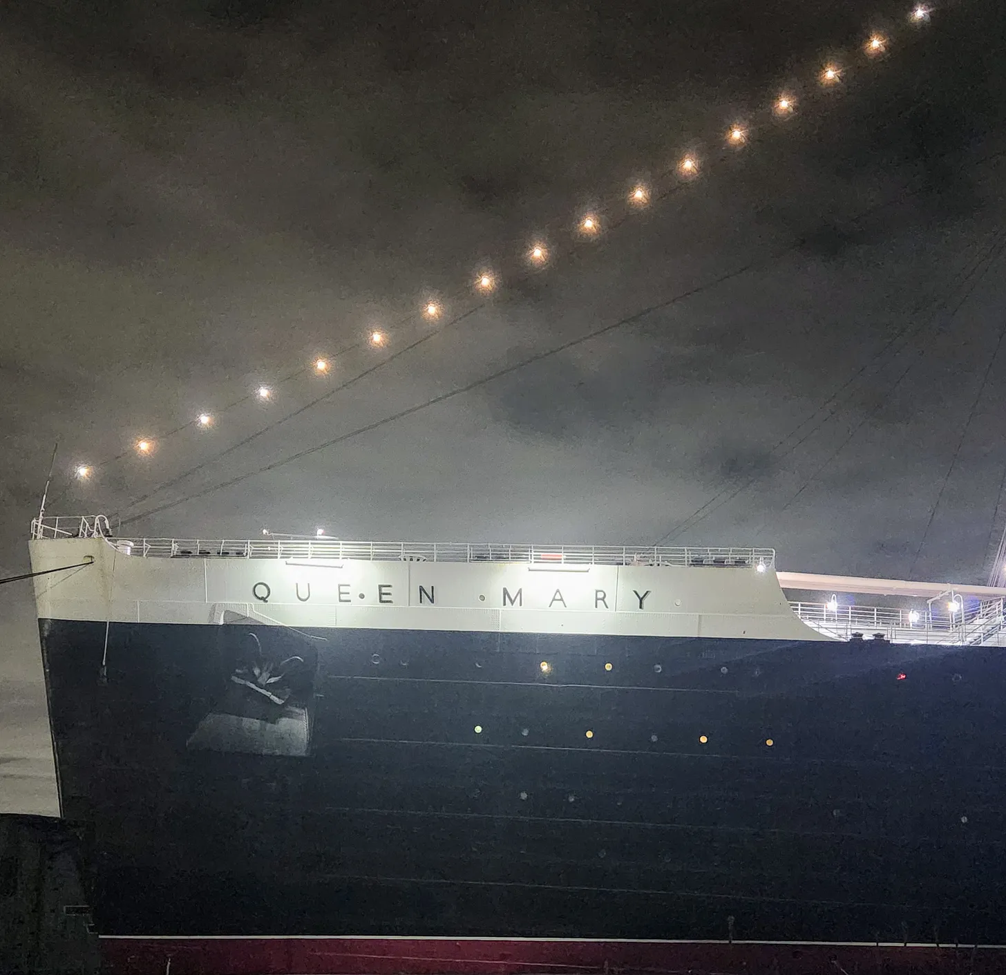 The bow of the RMS Queen Mary, seen from the side at night. The ship's hull is black with a white stripe at the top, on which her name is written in simple black letters.