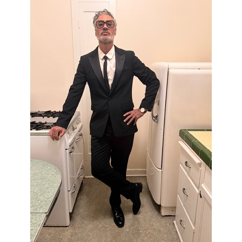 Taika Waititi stands in a small kitchen, dressed in a black suit, a white shirt and black tie and polished black shoes. He is wearing glasses with large, suqare-shaped lenses and a thick frame, and he is looking directly at the camera with his head tilted back a little, chin raised. He is leaning on a gas stove to his side with one hand, his other hand placed on his hip, and he has one leg bent at the knee to cross it over his other leg, bringing the toe of his shoe to the floor with the sole facing outwards.