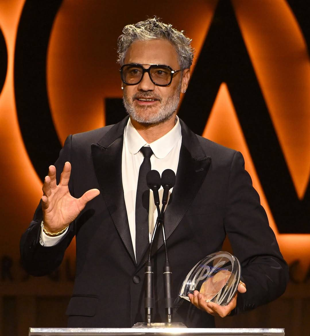 Taika speaks at the PGA Awards ceremony, holding his award in one hand and gesturing with the other. He is looking slightly off to the side and smiling as he talks.