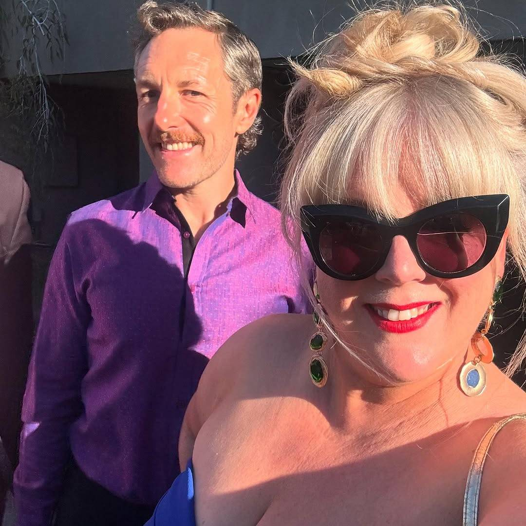 Selfie of Gypsy and 'The Newsreader' writer Michael Lucas, both looking at the camera with open-mouthed smiles. Gypsy is in the foreground, wearing large cat-eye sunglasses, red lipstick, long dangly earrings and a blue strapless dress, her blond hair in a messy top knot. Michael stands behind her, dressed in a purple button-up shirt and black trousers.