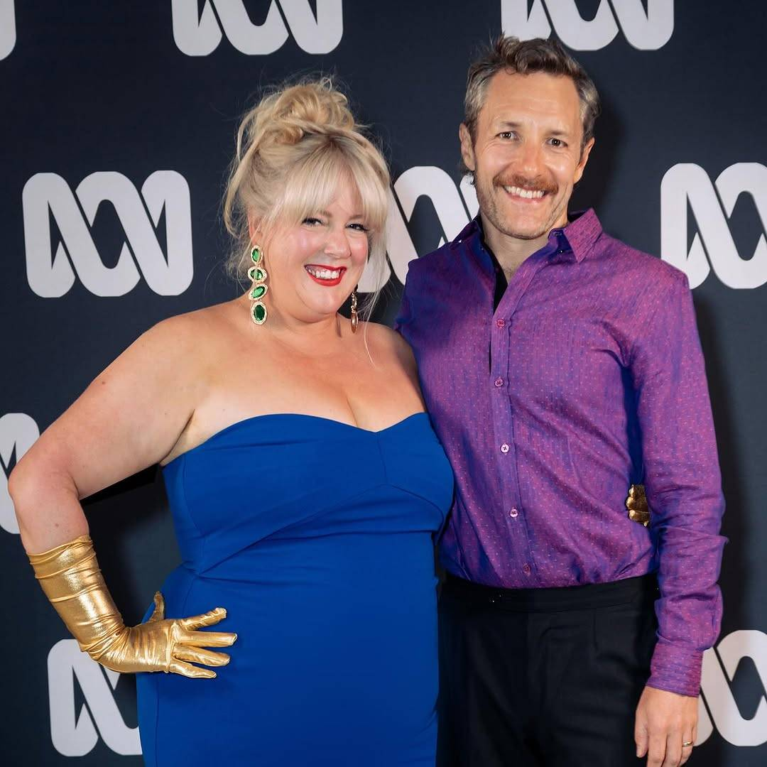 Gypsy Taylor and Michael Lucas stand shoulder-to-shoulder as they pose for press photographers at the season premiere. Both are looking at the camera with open-mouthed smiles. They are seen from about mid-thigh up so we can see more of their outfits. Gypsy has one hand placed on her hip, showing off her golden forearm-length gloves.