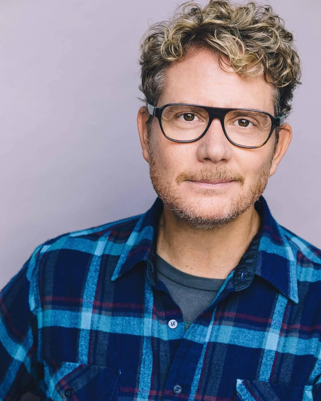 Head shot of Brian Gattas. Brian is looking at the camera with a warm closed-mouth smile. He has short curly hair that is blond at the tips and black at the roots, with a bit of grey coming through at his temples. His short beard and mustache are a mix of blond and grey, and his eyes are brown. He wears glasses with a black rim that is thin around the lenses but thick at the bridge. He is wearing in a blue flannel shirt with a grey t-shirt underneath.