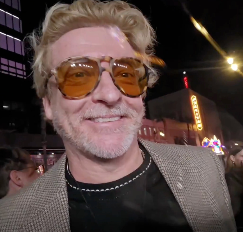 Screencap from a video posted on Rhys's Substack page. It is a slightly blurry shot of him outside the Love Hurts premiere venue, smiling at someone or something just to the side of the camera.