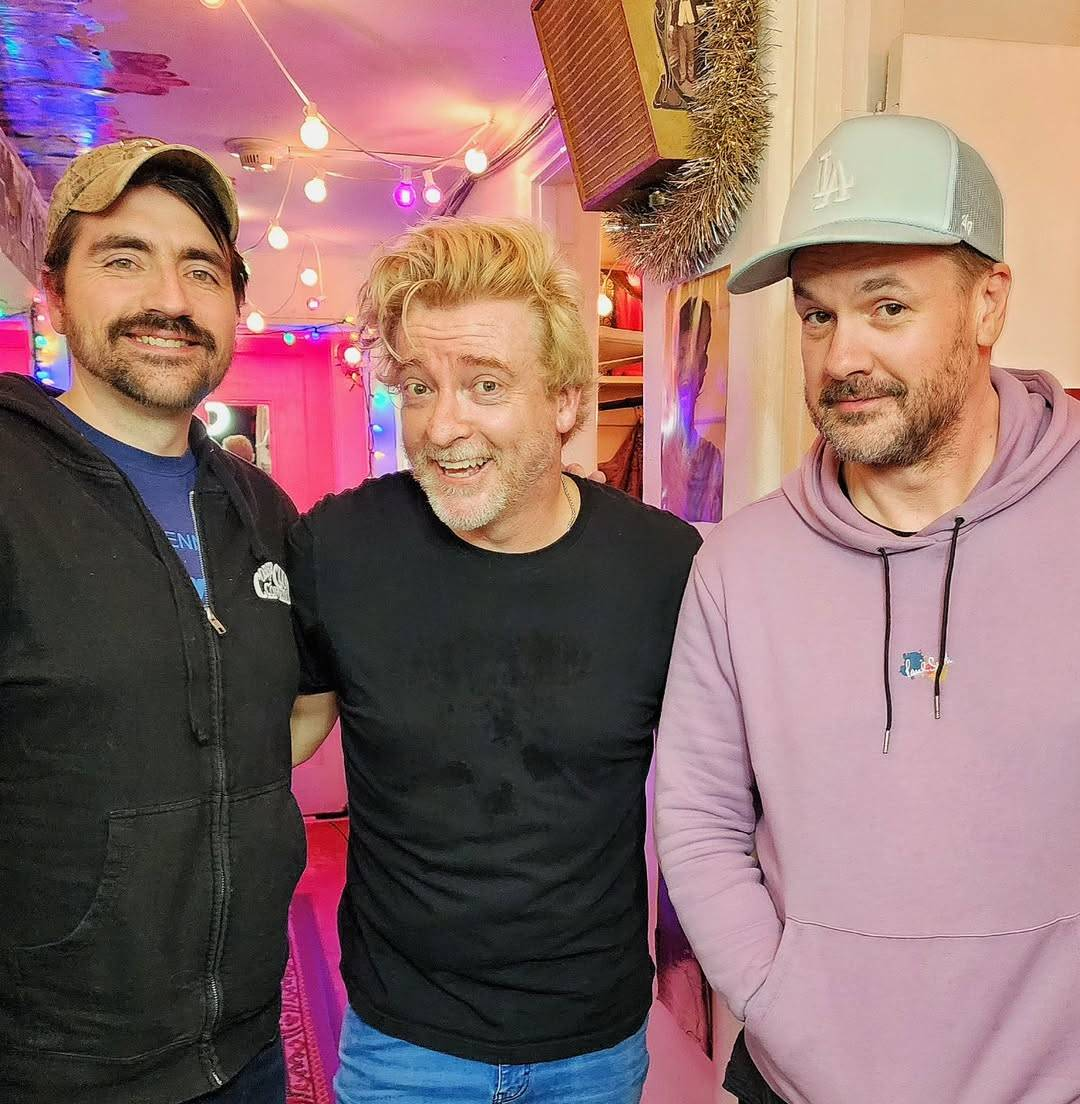 Photo of Rhys with Jim Jefferies and Trae Crowder at the Largo in Los Angeles. They stand inside a brightly lit corridor with Rhys in the middle, Trae on his left and Jim on his right, all 3 of them smiling at the camera.