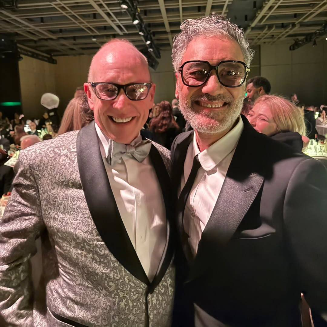 Taika Waititi and Kevin Goetz at the PGA Awards. They are seen from the waist up, standing close in a crowded room. Both are smiling broadly, and Kevin is looking at the camera while Taika is looking at something or someone just to the side.