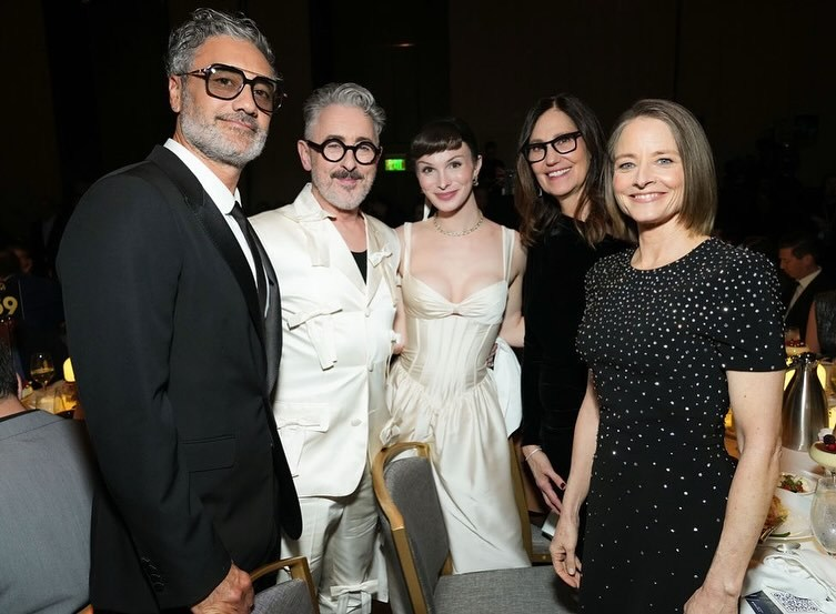 Taika Waititi, Alan Cumming, Dylan Mulvaney, Mari Jo Winkler and Jodie Foster at the PGA Awards. They all smile for the camera while standing in a kind of semi-circle, squeezed a bit awkwardly into the tight spaces between chairs and tables.