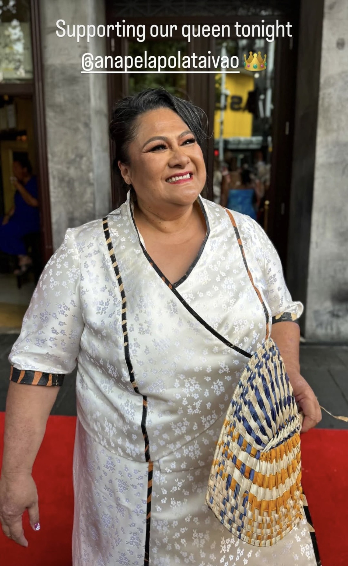 Photo of Anapela on a red carpet outside the premiere venue, looking at someone just off to the side with a big smile on her face. She is wearing her hair in an updo, and she is dressed in a lavalava and a matching wrap top, both made from a shiny silvery fabric embroidered with clusters of small flowers. She is holding a Samoan woven hand fan in one hand. Text at the top of the photo reads: 'Supporting our queen tonight @ AnapelaPolataivao'.Photo of Anapela on a red carpet outside the premiere venue, looking at someone just off to the side with a big smile on her face. She is wearing her hair in an updo, and she is dressed in a lavalava and a matching wrap top, both made from a shiny silvery fabric embroidered with clusters of small flowers. She is holding a Samoan woven hand fan in one hand. Text at the top of the photo reads: 'Supporting our queen tonight @ AnapelaPolataivao (Crown emoj)'.