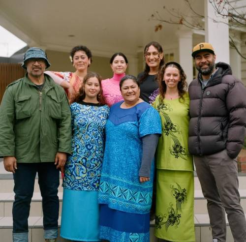 Anapela and 7 film crew members smile at the camera as they stand on some steps outside a building. Anapela and the other 5 women, who were all part of the costume team, are dressed in puletasi (a traditional Polynesian outfit consisting of a skirt and a long top) of different colours and patterns. 