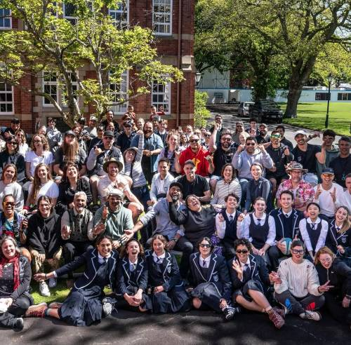 Group photo of cast and crew members from the film sitting or standing outside a red brick building on what looks like a school campus. Anapela is laughing, sitting in the second row from the front amidst a group of young actors in school uniforms.