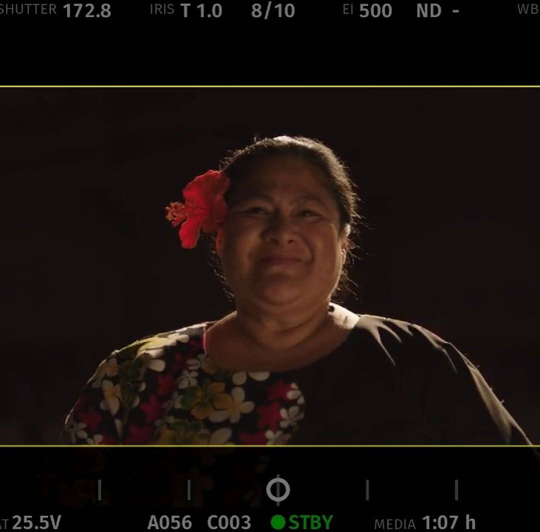 Picture of a camera screen showing a close-up of Anapela in character, shot head-on. She is smiling proudly as she looks up at something beyond the camera. She is wearing a red hibiscus flower in her hair, which is pulled back, and a black top with a multi-coloured flower pattern. 