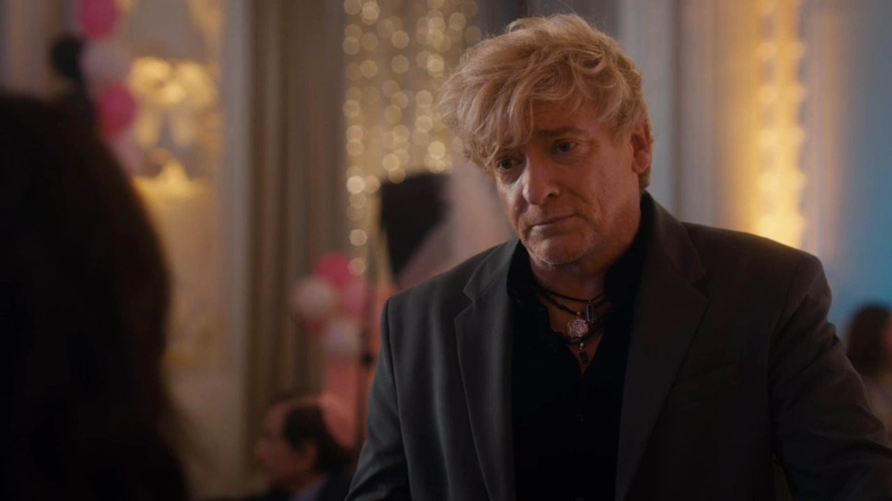 Rhys in 'The Z-Suite'. His character has tousled blond hair that is longer on top and shorter at the sides and he is looking down at someone out of frame with his eyebrows drawn together and his lips pressed together in a straight line. He is wearing a grey suit jacket that looks a bit too large around the shoulders, and a black shirt with the top few buttons open to reveal several leather necklaces.