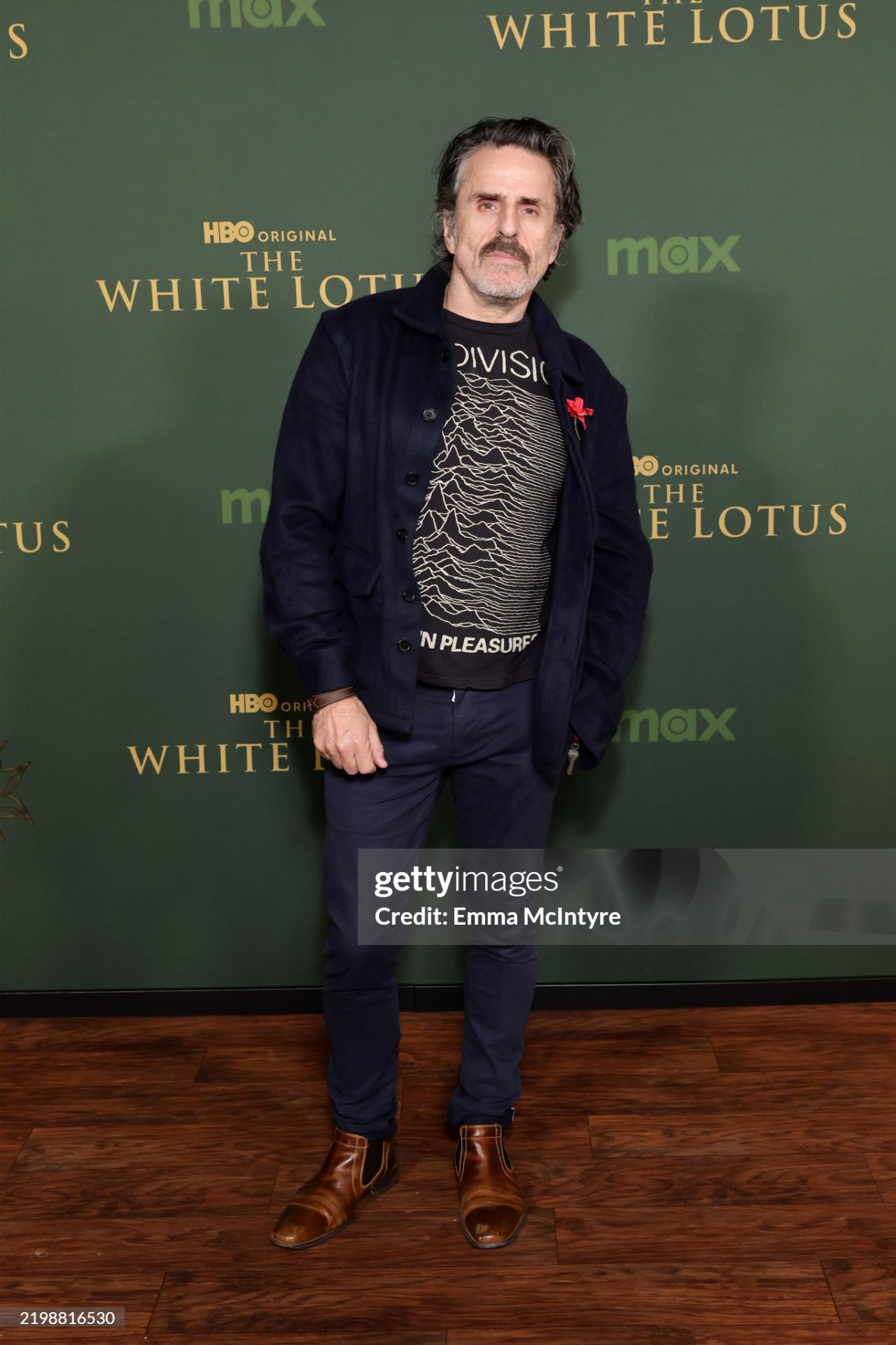 Con stands in front of a dark green wall inside the series premiere venue. He is looking at the camera with one corner of his mouth slightly quirked up, dressed casually in a Joy Division t-shirt paired with a thin dark blue jacket, blue trousers and brown Chelsea boots. A red flower is pinned to his jacket over his heart.