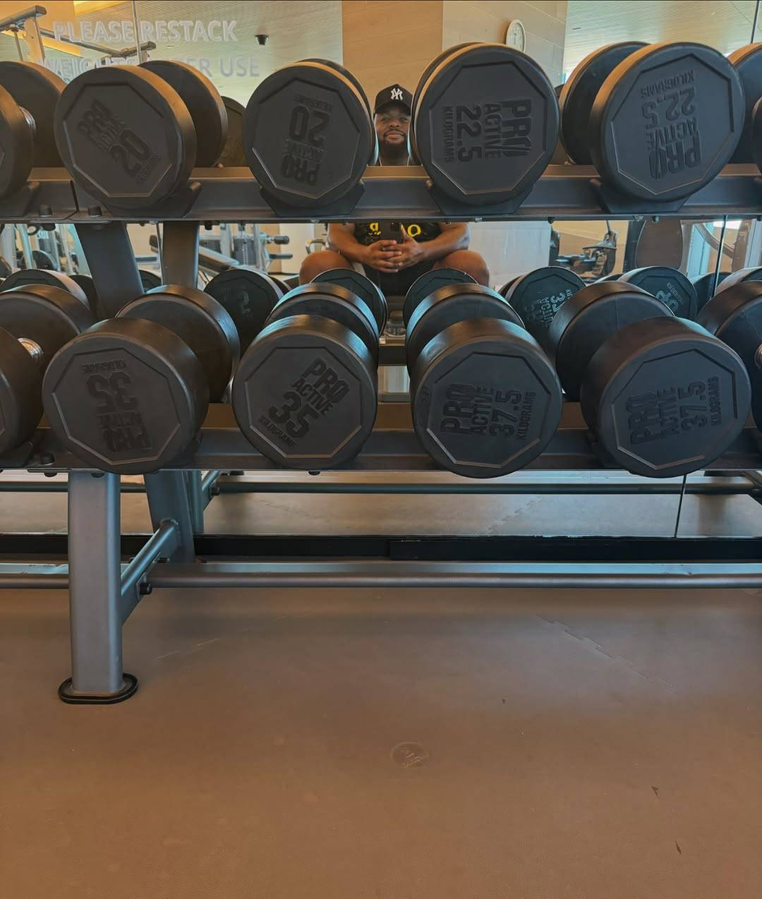 Photo of Samson's reflection in a mirror behind a rack of dumbbells in a gym. He is sitting on a bench in front of the rack and looking at the mirror on the wall behind it with a small smile, holding his phone in both hands at hip level to take the picture.