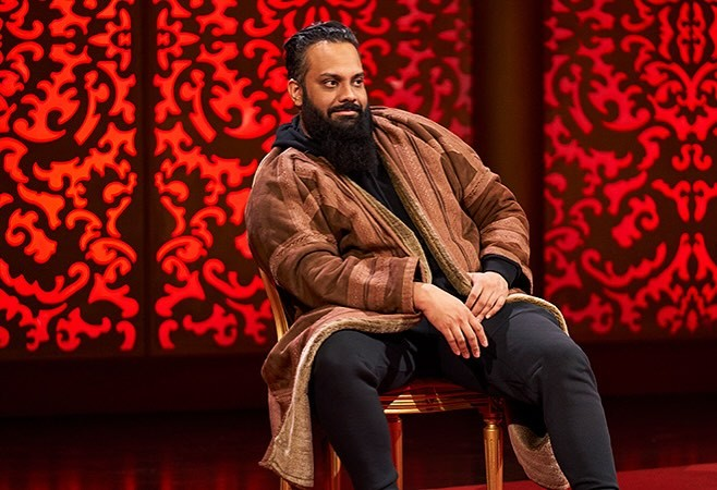 Guz Khan sits on the Taskmaster stage. He is looking at something out of frame with a slight smile, one elbow leaning casually on his leg. He is wearing a soft-looking brown coat over a black hoodie and black trousers.