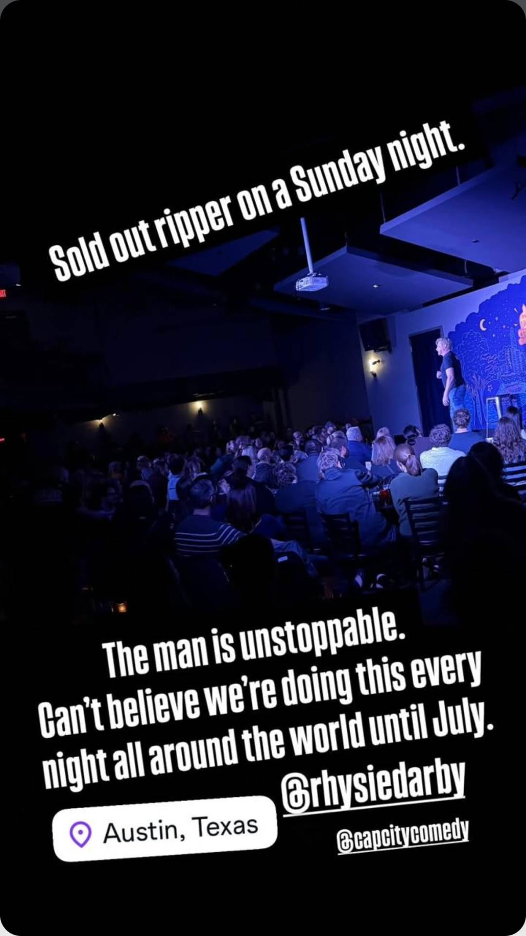 Photo from one of Rhys's stand up shows, seen from the back of the audience. Rhys is standing on stage and facing away from the camera. Text reads: 'Sold out ripper on a Sunday night. The man is unstoppable. Can't believe we're doing this every night all around the world until July.' @ RhysieDarby and @ CapCityComedy are tagged at the bottom and there is a location tag for Austin, Texas.