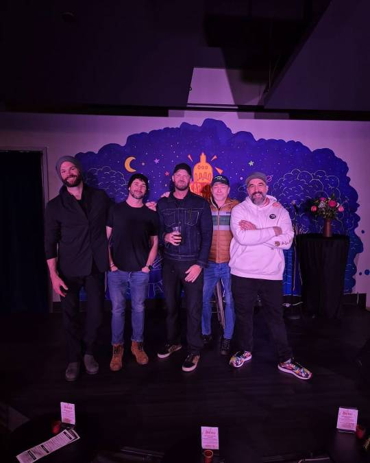 Rhys stands on stage next to Jared Padalecki, Steve Wrigley and two other men, all of them smiling at the camera.