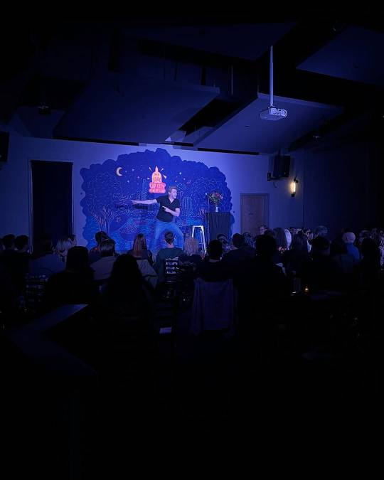 Another photo taken from the back of the room at Rhys's Cap City show. Rhys is standing on stage and pointing to one side with both hands, one arm outstretched and the other bent at the elbow, while looking in the opposite direction.