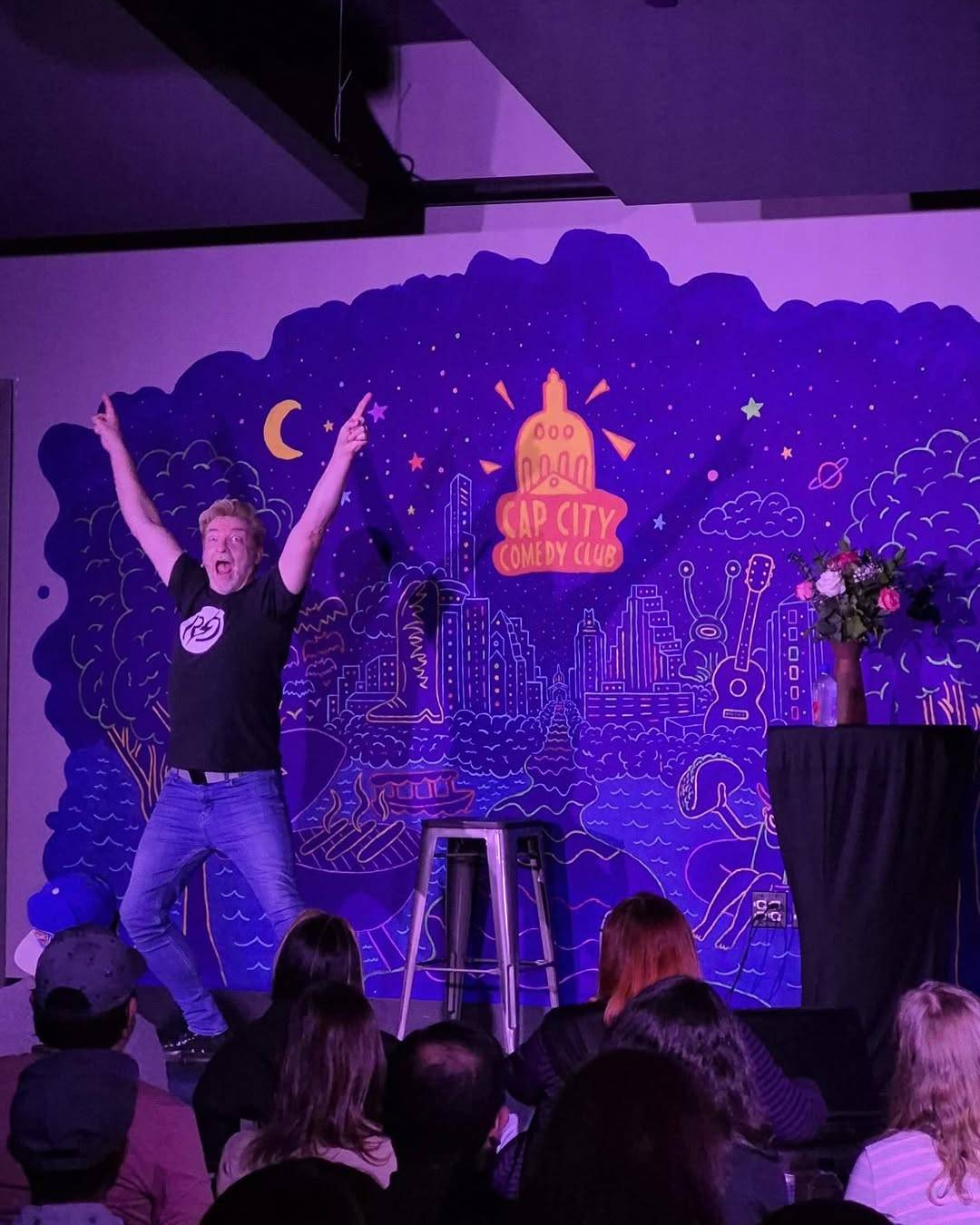 Rhys on stage at Cap City, this time seen from closer to the stage. He is standing with both hands in the air, index fingers pointing upwards, his eyes wide and mouth open.