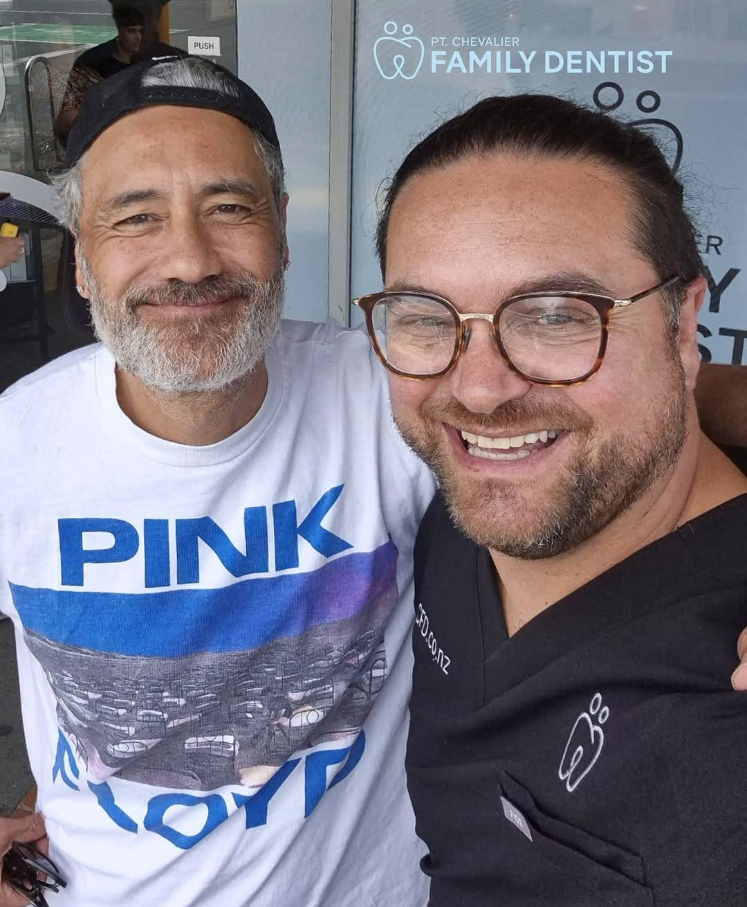 Taika and his dentist stand in front of a glass door with ‘Pt. Chevalier Family Dentist’ written on it. They are both smiling broadly at the camera and Taika has one arm around the other man’s shoulder.