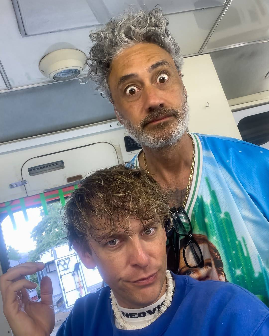 Photo of Taika and a young man inside what looks like a trailer on a film set. Taika is staring at the camera with wide eyes, his brows raised and his lips in a straight line. The other man is sitting in front of him, also looking at the camera but with a skeptical expression, his lips slightly pursed and one corner of his mouth quirked up.