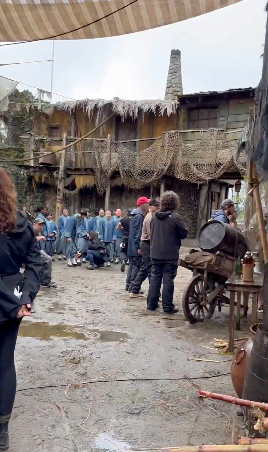 Still from a behind-the-scenes video posted by Vico. It shows part of the Republic of Pirates set from Our Flag Means Death season 2, with members of the film crew working in the foreground while the actors playing Zheng’s crew wait in the background.