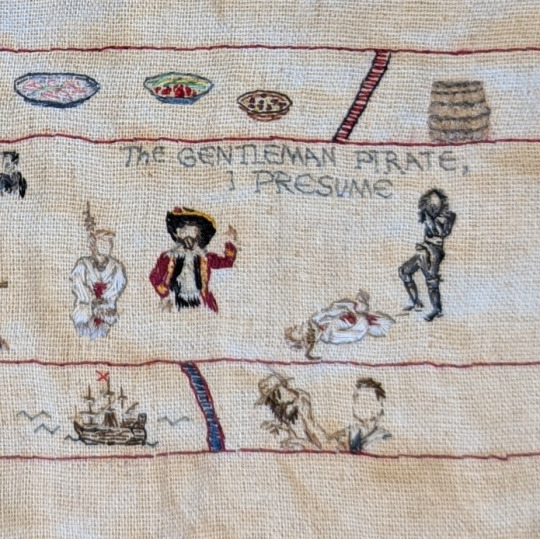 Close up photo: Stede hanging from a noose after getting gut-stabbed, the Spanish captain in his red coat and big hat, and the meet-cute scene, with Stede lying bloodied on the floor as Blackbeard looks down at him. The line 'The Gentleman Pirate, I presume' is embroidered at the top.