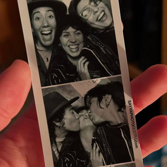 Two photos on a strip of black & white photobooth pictures of Vico and their partners, held in Vico's hand. In the photo at the top, all 3 of them are laughing, and the bottom pic shows them kissing. 